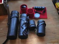 Four black leather lens cases with straps laid out on wooden surface, small accessory items and red interior of carrying box partially visible outside in background