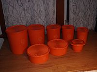 All 8 orange Tupperware containers arranged on a wooden table, showing various sizes with intact lids in a bright orange color and floral emblem decoration.
