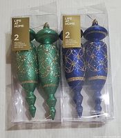 Two sets of holiday ornaments in clear boxes, one set green and one set blue, each set containing two ornaments decorated with gold glitter patterns.
