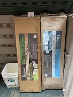 Photo showing two unopened boxes of galvanized metal raised garden beds with labeling and one white plastic bucket beside them.
