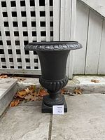 Side view of the black cast iron garden urn on a stone patio next to a white lattice fence showing the urn's decorative rim and base.