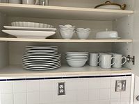 Shelves showing stacked white ceramic plates, bowls, mugs, and serving pieces including fluted pottery and covered butter dish.