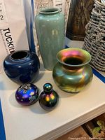 Five artisan signed vases and decorative glass and pottery items displayed together on a white tabletop against a mixed background.