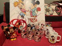 Group photo of all collectibles on red surface including Mickey & Minnie glass mugs, pitcher, toy banks, rolling toy, and poster in background