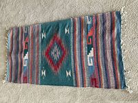 Full view of the large wool kilim rug showing central diamond geometric pattern in teal, red, and white, with surrounding striped multicolor bands and knotted fringe tassels on the short edge.