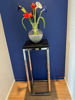 Full view of the plant stand with vase on top against a blue wall and light wood floor.