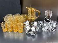 Full lot photo showing amber pitcher and glasses, clear floral glasses, and penguin/puffin glass dispensers and salt & pepper shakers