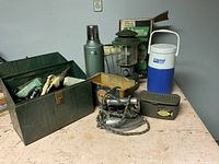 Photo of the full lot including metal box with fishing gear, vintage electric iron, Coleman lantern, Stanley thermos, and insulated beverage holder, arranged on a table.