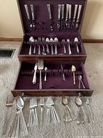 Full view of wooden case holding diverse assortment of silver-plated flatware including spoons, forks, knives, and serving utensils, laid out on purple velvet lining. Some flatware is outside the case.