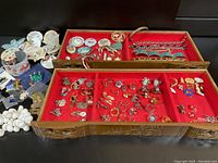 Wide photo of assorted vintage jewelry in three-section wooden tray lined with red velvet, displaying rings, brooches, bracelets, and pins including porcelain painted items and magnetic therapy bracelets.
