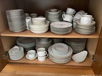 Dishes stacked on shelves, various sized plates, mugs, and cups, predominantly white with some gold rim plates visible
