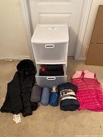 Full view of lot showing black down jacket, pink vest, rolled down blankets, and white plastic drawer bin with folded clothing inside.