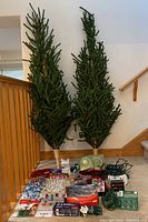 Two slim artificial Christmas trees leaning against stair railing with multiple assorted Christmas decorations arranged in front including lights, ornaments, garlands, and boxed items.