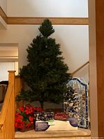 Front view of the unassembled 7.5 foot artificial Christmas tree on a stand with boxed light strings, garland, and red poinsettia basket at the base. Snowman LED light display visible boxed on right.