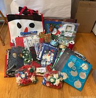 Multiple Christmas gift bags, gift wrap rolls, bows, and ribbons laid out on wooden floor.