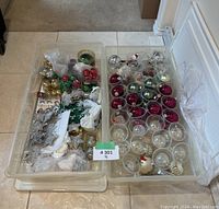 Two clear Rubbermaid plastic storage bins containing assorted Christmas ornaments including red, green, silver balls, snowflakes, and star shapes.