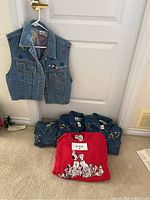 Photo showing hung sleeveless denim vest with embroidered Mickey Mouse and heart details, three folded denim button shirts with Mickey Mouse embroidery, and one red sweatshirt with 101 Dalmatians graphic.
