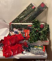 Photo showing boxed holly garlands, red fabric cloth, silver tinsel, packaged decoration items, and light strands all arranged together on surface.