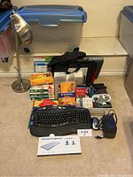 Photo of lot showing a silver adjustable desk lamp, assorted office supplies including boxed Bluetooth keyboard, black wired keyboard and mouse, printer paper, Vivitar DVDs and cartridges, power adapter, black gloves, and plastic storage totes.