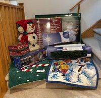 Photo showing entire lot arranged on carpeted stairs. Visible are the snowman figure, boxed spiral tree sculpture, boxed pine garlands, boxed string light sets, and two holiday rugs.