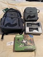 Photo showing the grey and black Roots branded backpack and smaller matching bag, boxed drinkware set, and outdoor adventure kit box arranged on a bed