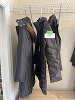Three black down jackets hanging on a rack including a long coat with faux fur trim on hood, a black vest, and another long coat.