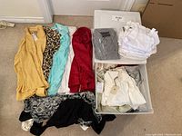 Lot of women's dress shirts folded and stacked on carpet with bins containing more shirts in background.