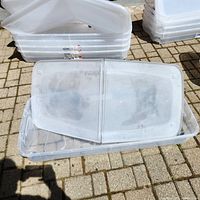 Clear rectangular plastic storage bin with bi-fold lid open, placed outdoors on paved surface, showing semi-transparent lid and container base.