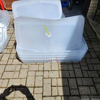 Four stacked clear plastic storage bins with bi-fold lids and visible casters underneath, photographed outdoors on stone pavers.