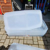 Four stacked clear plastic bins with bi-fold lids laying on top, outside on a paved surface.