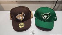 Front view of two Toronto Blue Jays caps, one brown with fur-lined ear flaps and one green with standard baseball cap design.