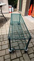 Green metal wire shelf unit with two shelves on paved outdoor surface, showing the structure and caster wheels.