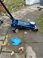 Blue 3 ton hydraulic floor jack on stone patio next to a single blue glove, showing side view with label and wheels visible.