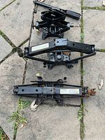 Three assorted black metal scissor jacks laid on stone pavement, showing varying sizes and designs with crank handles, visible wear, and usage stickers.