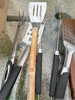 Close-up of four BBQ tools on glass surface: fork, slotted spatula with wooden handle, tongs with black handles, and another tool partially visible.