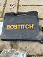 Black plastic carrying case with large Bostitch logo in yellow-orange letters placed on pavement ground with some leaves and soil marks around.