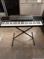 Full front view of Casio CTK-510 keyboard on black adjustable stand on tile floor. White cabinets background.