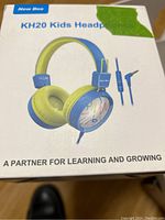 Front of box showing New Bee KH20 Kids Headphones in blue and yellow with colorful design on ear cups and wired connection.