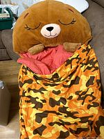 Closed view showing plush bear head pillow at top, camouflage outer layer fully zipped, sleeping sack laid out on couch.