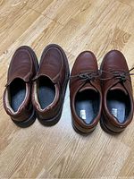 Two pairs of men's brown leather shoes side by side on a wooden floor. One pair is slip-on; the other has laces.
