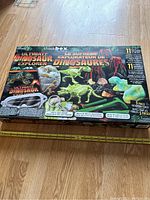Top view of the ThinkBox Ultimate Dinosaur Explorer kit box featuring dinosaur illustrations and description of 11 activities, 5 piece education tools, and colorful graphics.