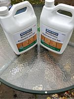 Two white jugs of Wood Shield Wood Restorer with readable product label showing usage for wood restoration and brightening. Jugs are unopened and placed on glass table.