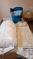 Full lot with white comforter on left, gold-patterned spread on right, two blankets in blue plastic tote behind