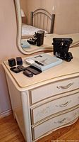 Overview of all items: binoculars with case, cell phone, weigh scale, and small items on a dresser.