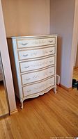 Front view of the tallboy dresser showing five drawers with floral designs and curved shapes, standing on curved legs on hardwood floor.