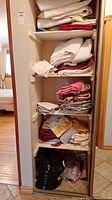 Shelving with stacked assorted linens including towels, tablecloths, and cloth bags at bottom