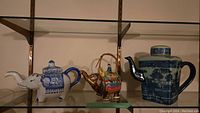 Three ceramic teapots on glass shelf: white and blue elephant shape, colorful elephant shape, large blue and white square teapot