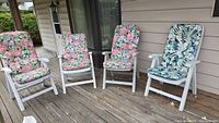 Photo showing four white PVC patio chairs on a wooden deck. Three chairs have pink and green floral padded cushions, one has a blue and green floral cushion.