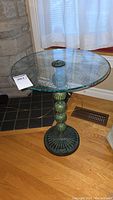 Side table standing on wood floor near stone fireplace. Shows metal base with ribbed and spherical details and round glass top with etched floral patterns.