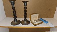 Wide shot of pair of twisted wood and sterling silver candlesticks standing side by side, set of small sterling spoons in a presentation case, and smaller box of spoons on shelf.